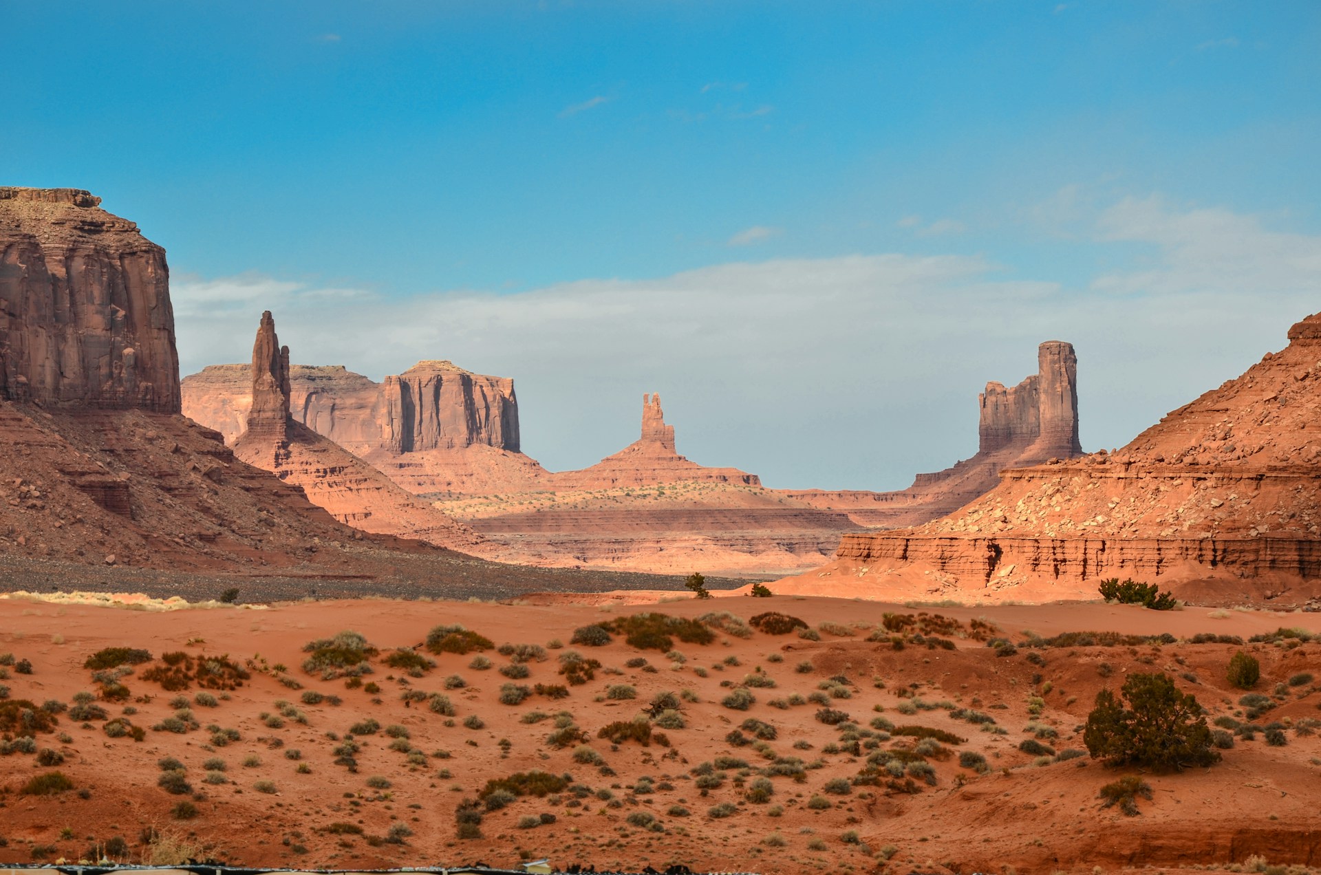 Panoramic view of red sandstone buttes and mesas in Monument Valley with desert shrubs in the foreground under a blue sky